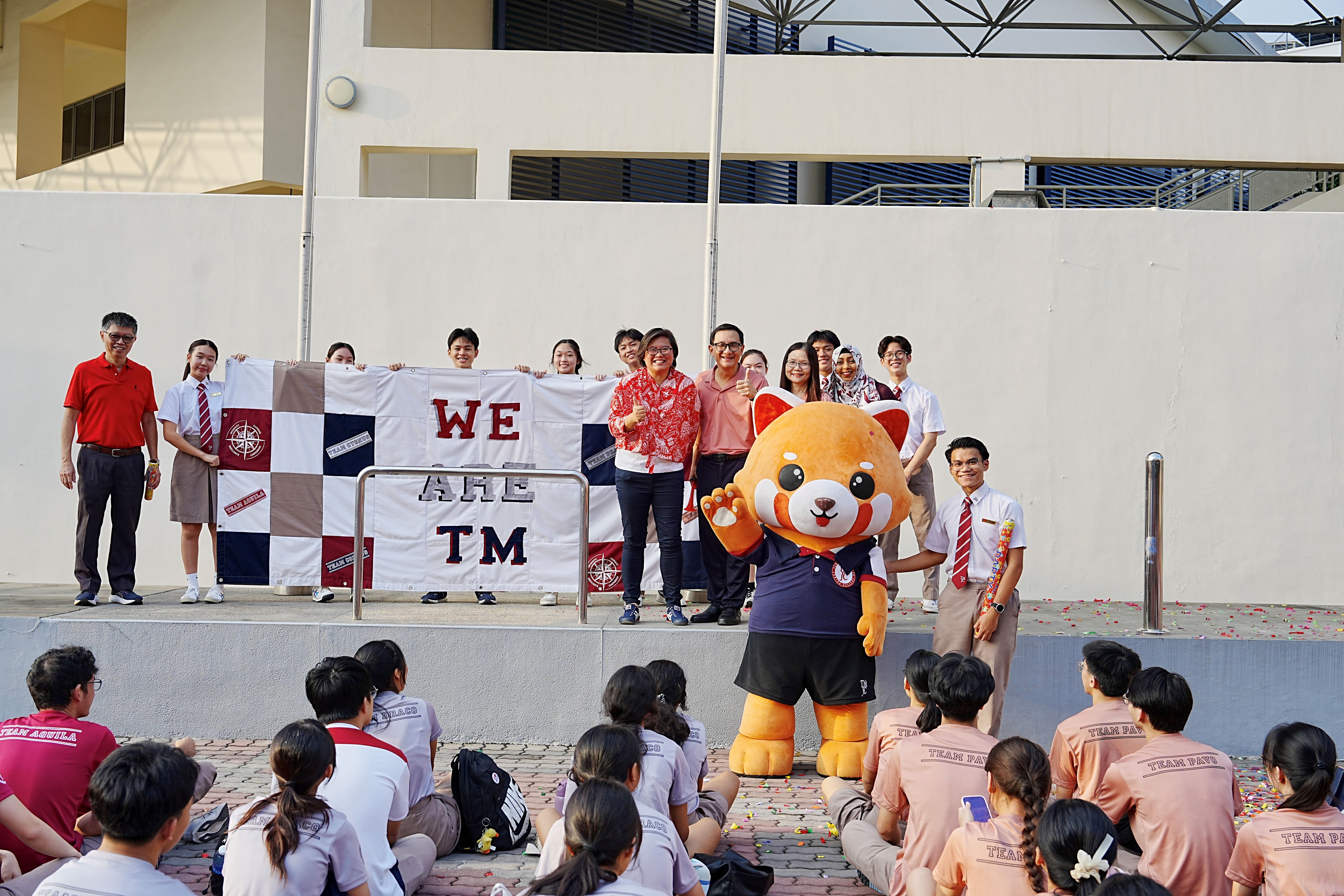 Students, teachers, and school leaders pose on a stage holding the completed "WE ARE TM" patchwork banner, with a red panda mascot in front and a seated student audience below.